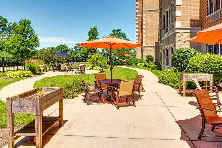 A sunny outdoor patio area with tables and chairs under an orange umbrella, surrounded by greenery and a building.