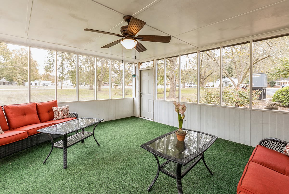A cozy sunroom featuring a ceiling fan, glass tables, and a green carpet with large windows offering an outdoor view.