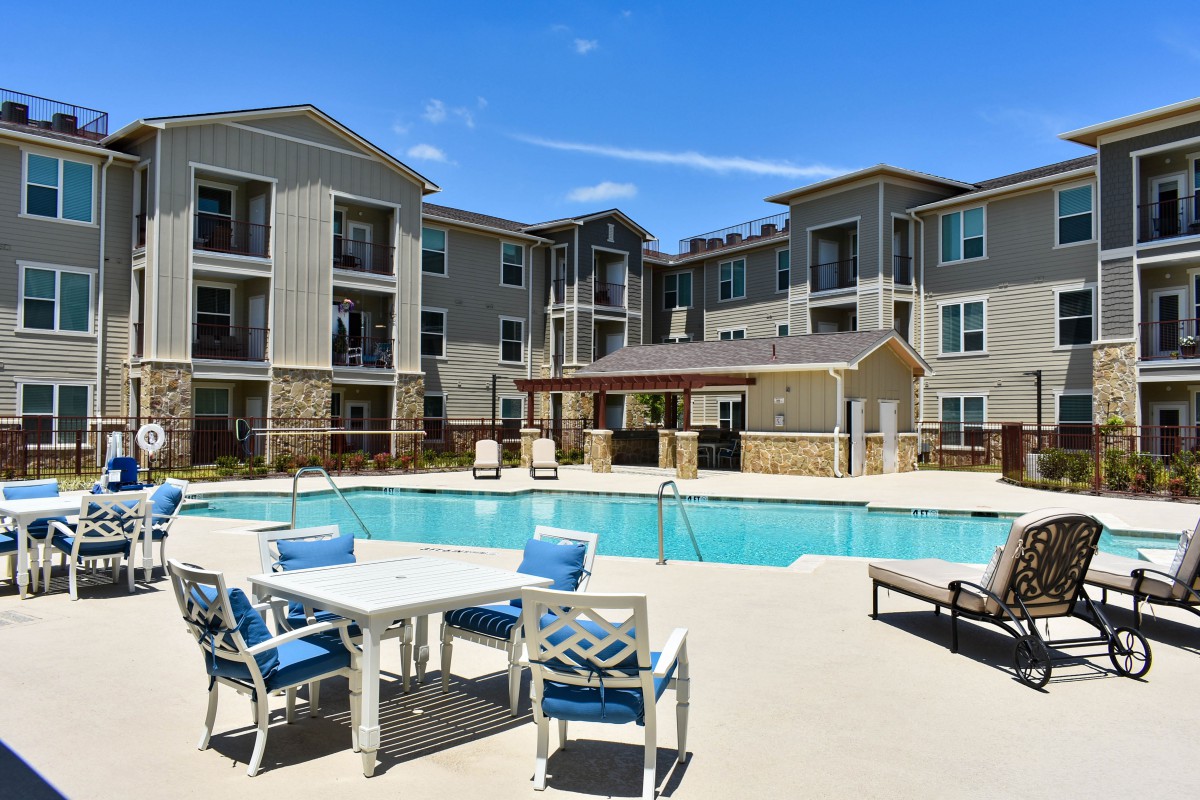 A modern apartment complex featuring a swimming pool and outdoor seating area under a clear blue sky.