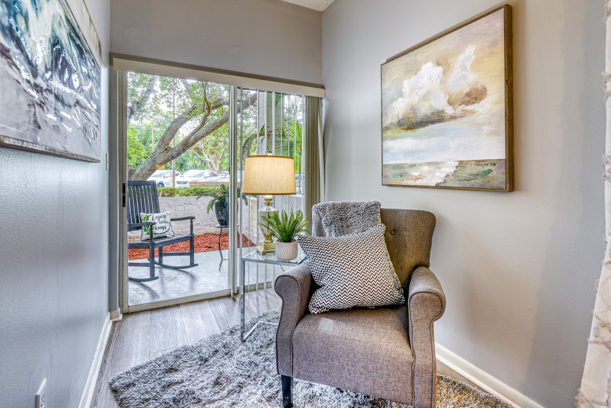 A cozy corner with an armchair, lamp, and a painting, overlooking a patio through large windows.