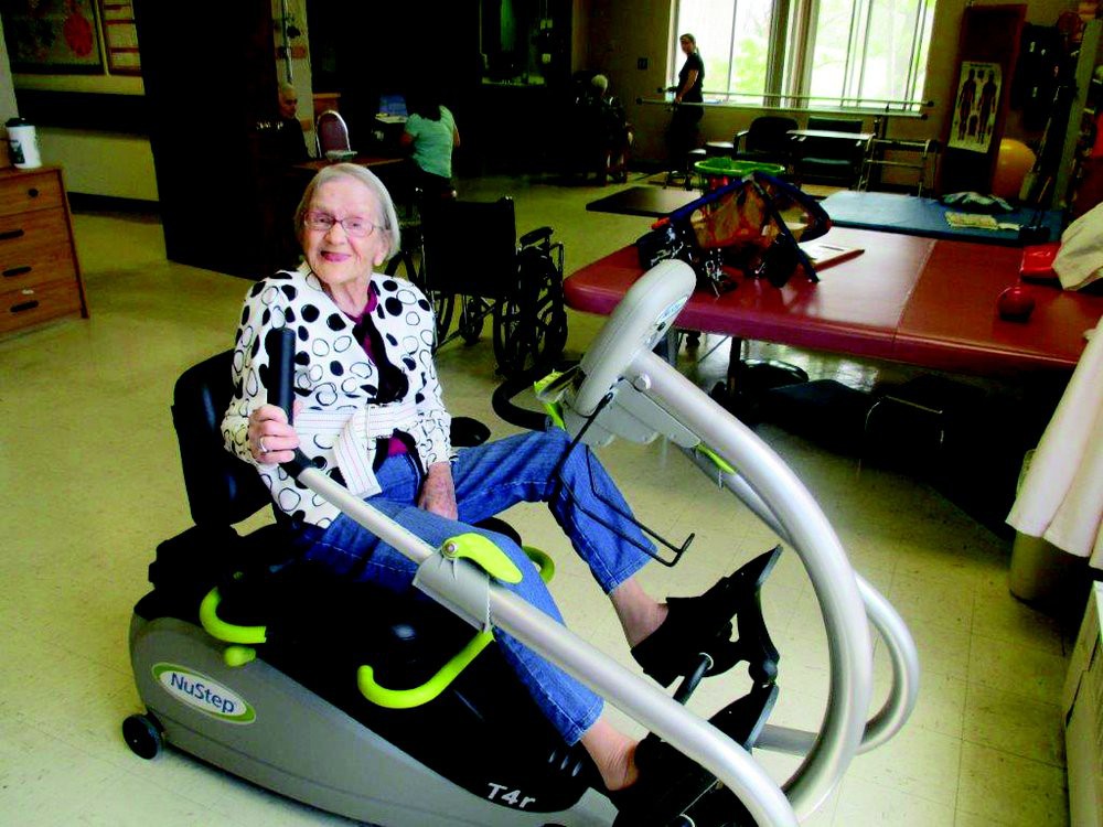 An elderly woman smiling while using exercise equipment in an indoor rehabilitation setting.