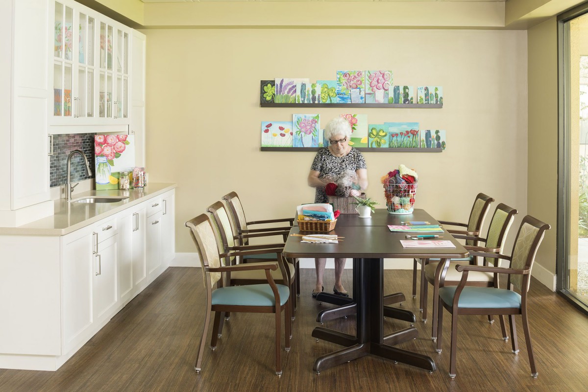 An elderly person stands in an art studio surrounded by colorful paintings and art supplies.