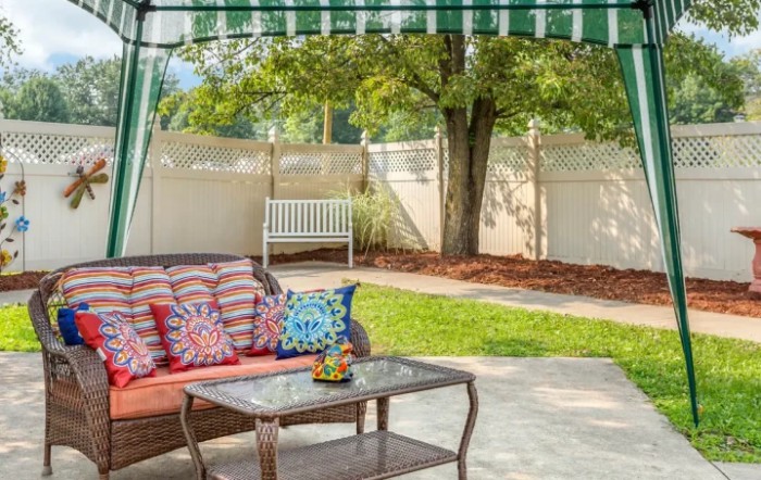 A cozy outdoor patio with a sofa, colorful pillows, and a table underneath an awning, surrounded by a garden with trees and a fence.