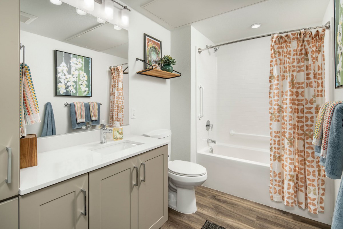A modern bathroom featuring a vanity with a large mirror, sink, and storage cabinets. It includes a shower with a patterned curtain and overhead lighting.