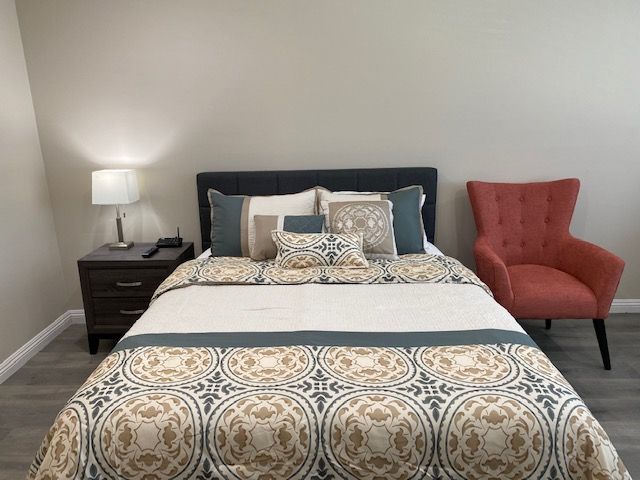 A neatly arranged bedroom featuring a bed with patterned bedding, a nightstand with a lamp, and an orange armchair.
