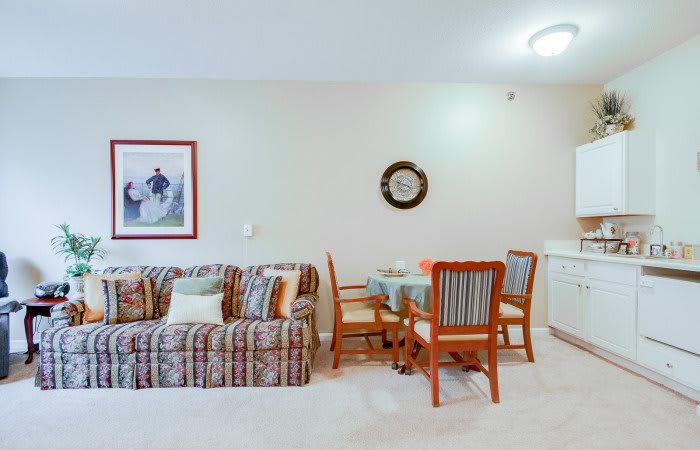 A cozy living room with a striped sofa, a small dining table with chairs, and decorative wall art and clock.