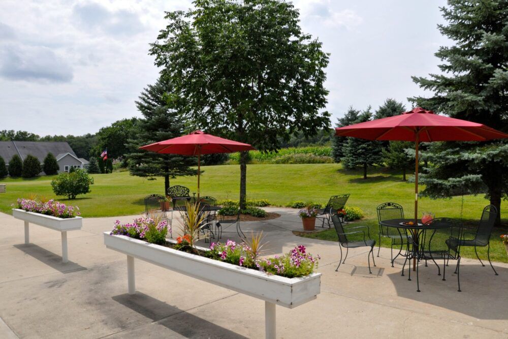 Outdoor patio area with tables and red umbrellas surrounded by a green garden and trees.