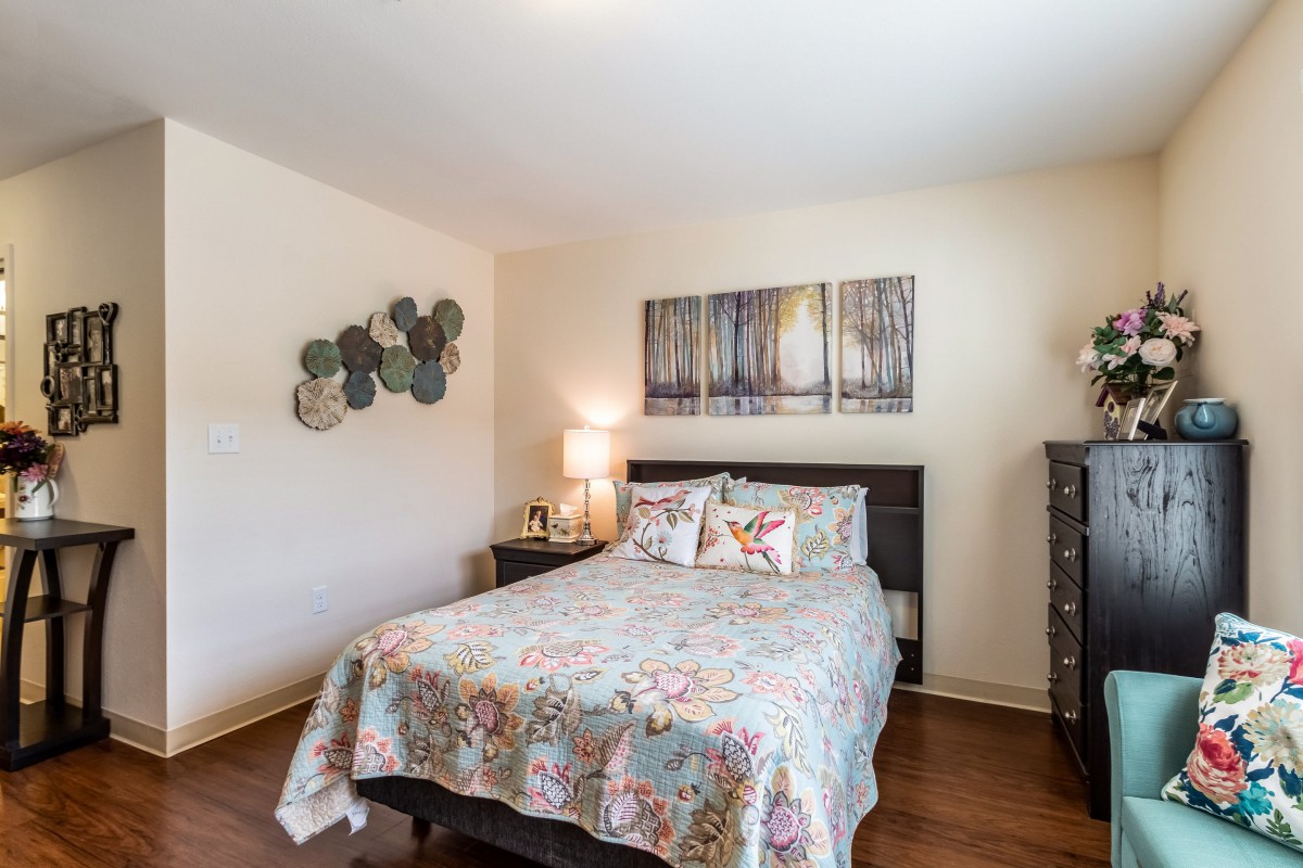 A cozy bedroom with a floral bedspread, artwork on the wall, and a warm lamp on the bedside table.
