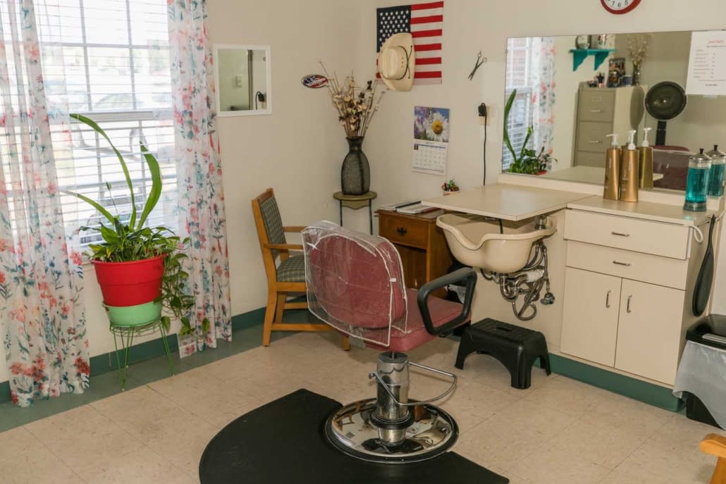 A vintage barber shop setup with a classic chair, mirror, and desk, featuring an American flag on the wall and plants by the window.