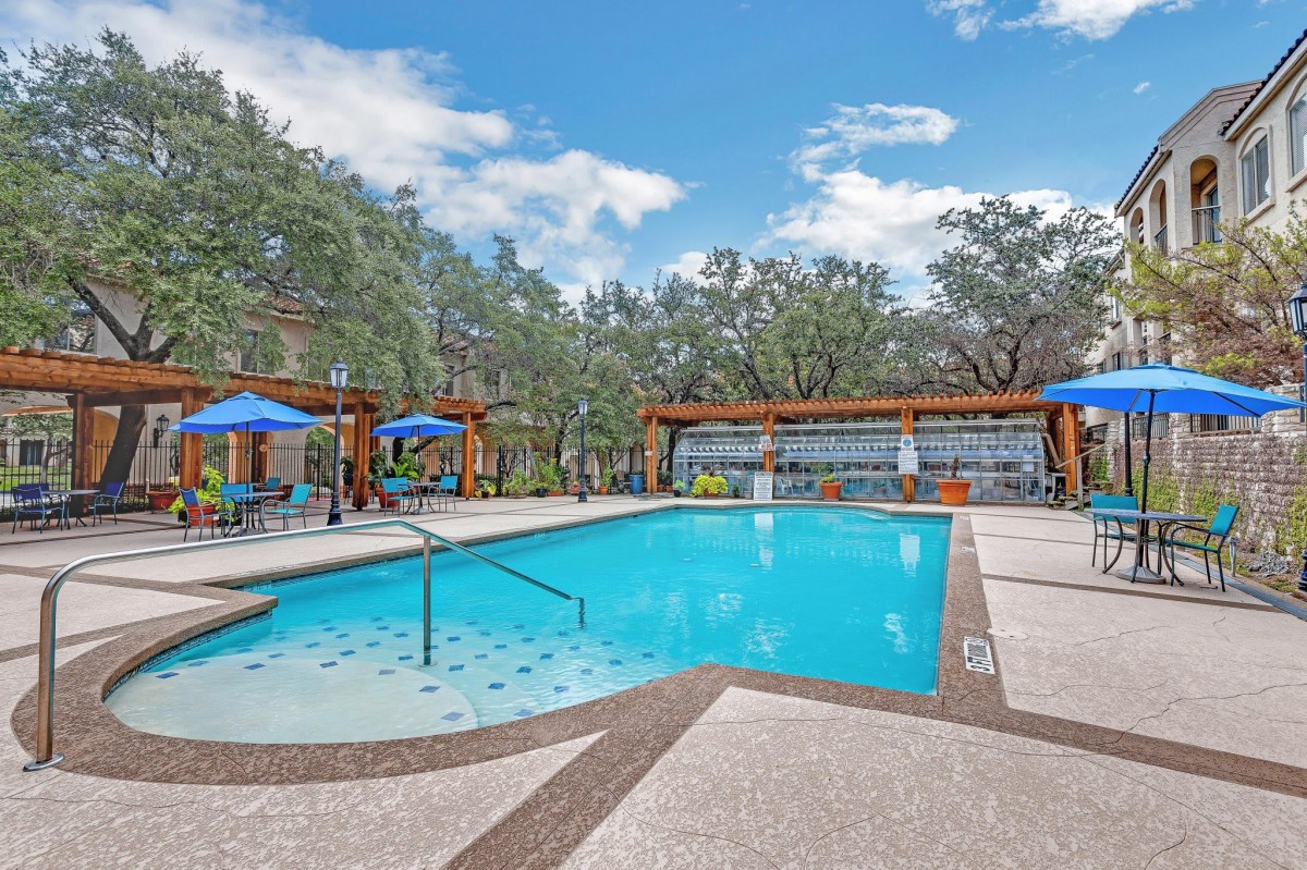 A serene outdoor swimming pool area with pavilions, umbrellas, and surrounding trees under a blue sky.