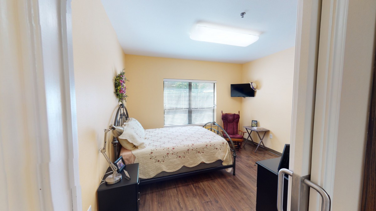 A cozy bedroom featuring a bed, nightstand, wall-mounted television, and a chair by the window.