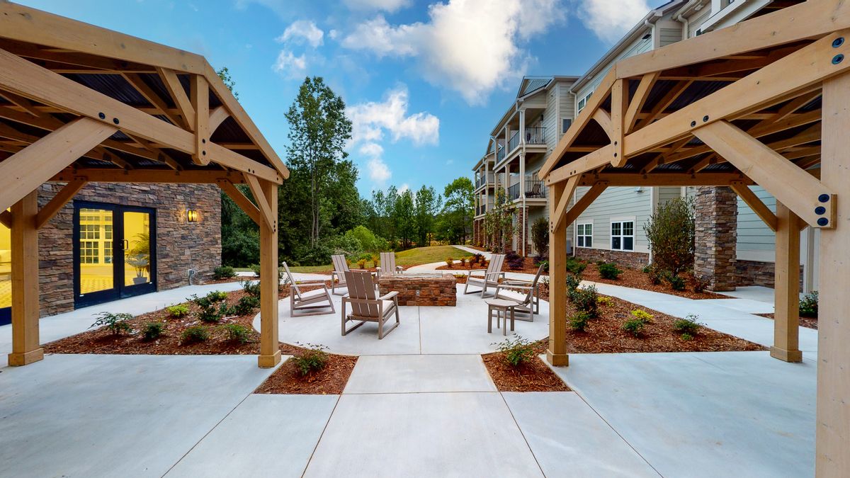 A modern outdoor patio area featuring a fire pit, surrounded by chairs and wooden gazebos, adjacent to an apartment building.