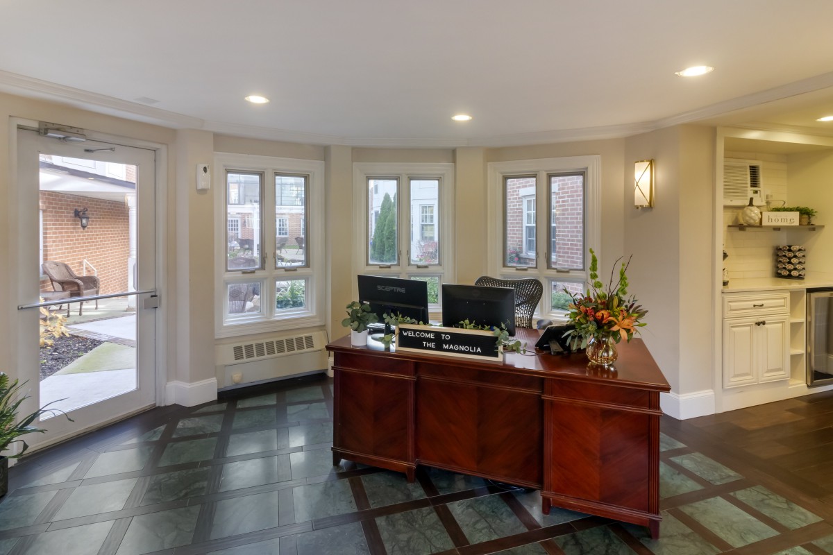 A well-lit office reception area featuring a wooden reception desk with computer monitors and floral decorations.