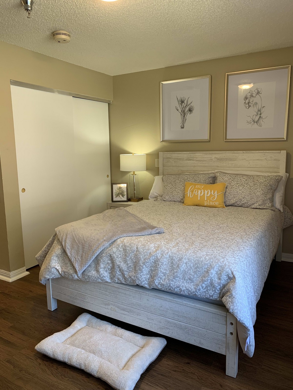 A cozy bedroom featuring a neatly made bed with decorative pillows, framed artwork above, and a side table with a lamp.