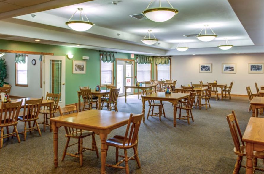 A spacious dining room with multiple tables and chairs, well-lit with ceiling lights and natural light from the windows.
