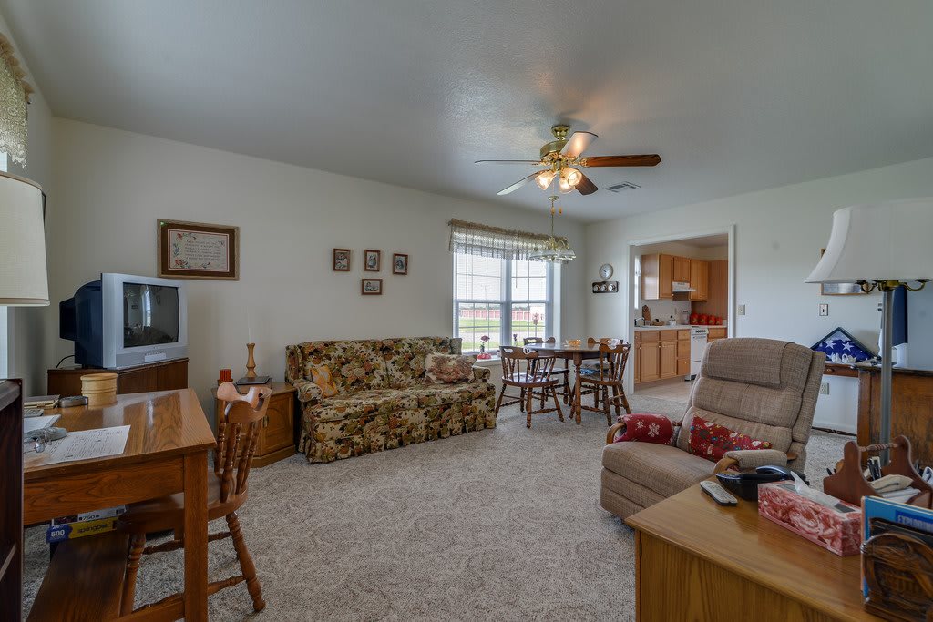 A cozy living room with vintage furniture, featuring a sofa, recliner, and a dining table near a window under a ceiling fan.