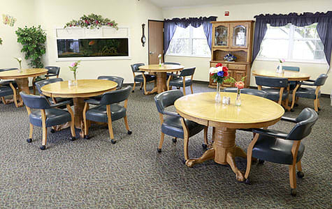 A cozy dining area with several round tables and chairs arranged on a carpeted floor, featuring windows with curtains and decorative accents.