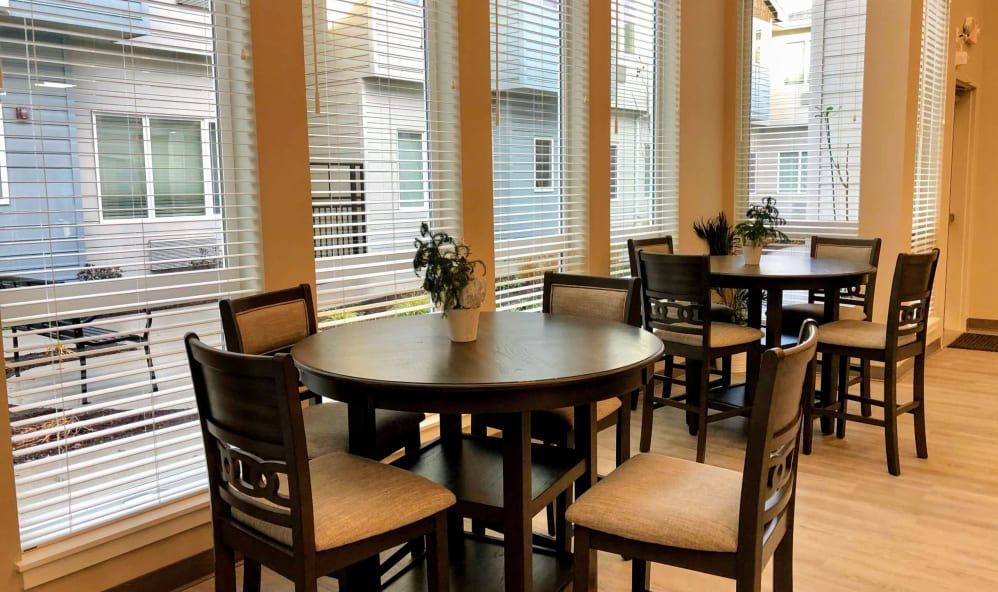 A cozy dining area with round tables and chairs, illuminated by natural light through large windows with blinds.