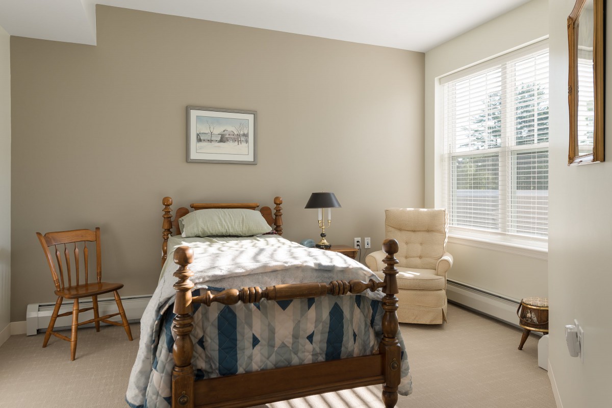 A cozy bedroom with a single wooden bed, an armchair, and a window letting in natural light.