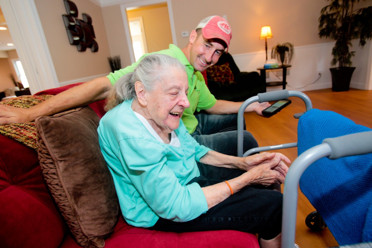 An elderly woman sits on a couch and smiles, accompanied by a man in a living room setting.