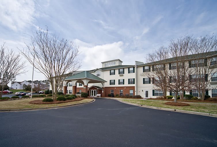 A large residential building with a paved driveway and surrounding trees under a cloudy sky.