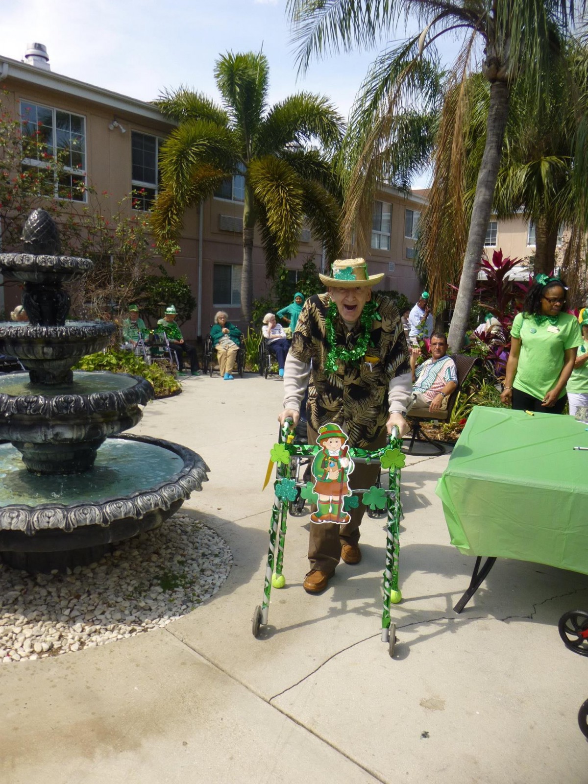 A person in festive green attire using a walker near a fountain, surrounded by tropical plants and other people.