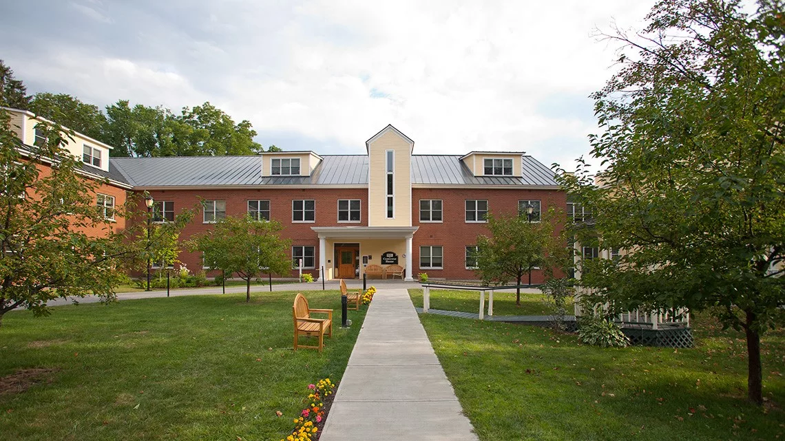 A brick building on a campus with a pathway and benches in the foreground, surrounded by trees.