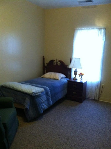 A cozy, simple bedroom featuring a single bed, a nightstand with a lamp, and natural light coming through the window.