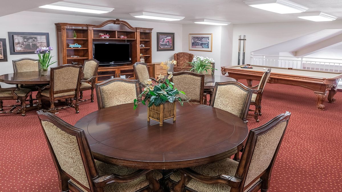 A spacious living room with a round wooden table and upholstered chairs, featuring a television, bookshelf, and plants for decoration.