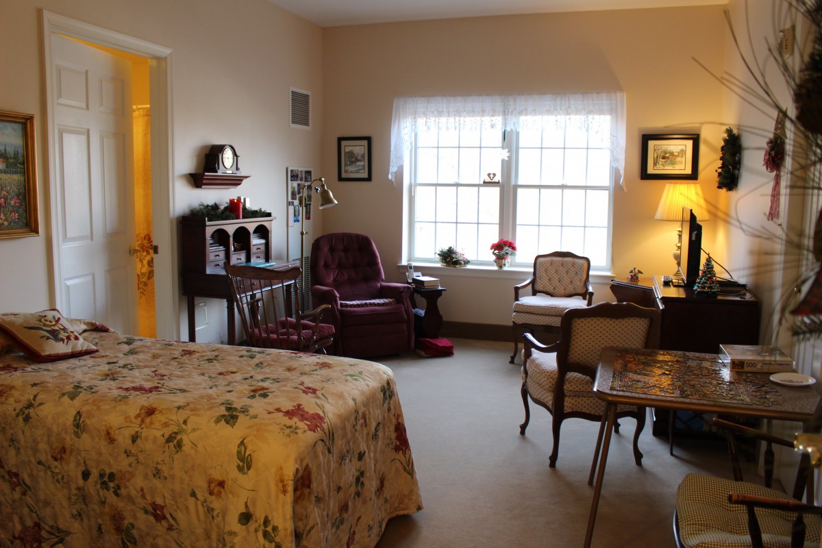 A cozy bedroom interior with traditional furnishings, featuring a bed, armchair, and a desk near a large window.