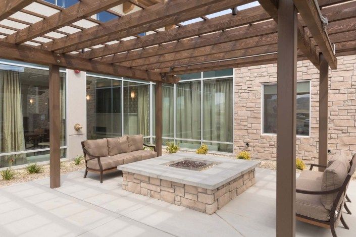 A modern patio featuring a wooden pergola, comfortable outdoor seating, and a central fire pit, surrounded by large glass windows and stone walls.