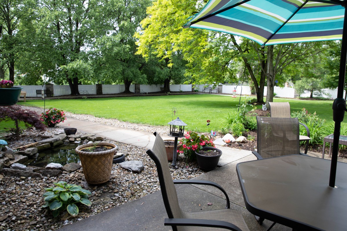 A peaceful backyard garden with a patio area, featuring chairs, an umbrella, and potted plants under lush green trees.