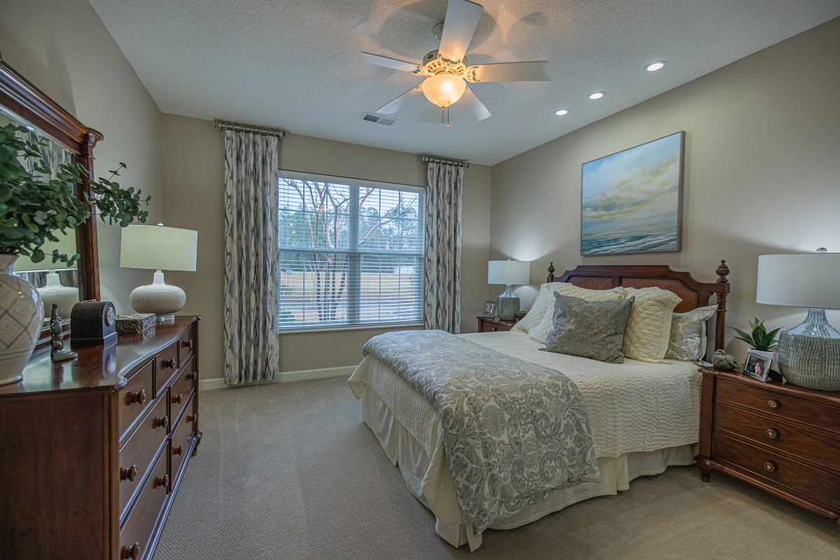 A cozy bedroom with a well-made bed, nightstands, and lamps, featuring a large window with curtains and a ceiling fan overhead.