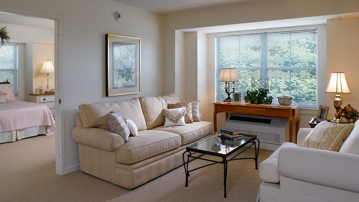A cozy living room with a beige sofa, glass coffee table, and a window letting in natural light.