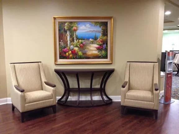 An elegant room interior featuring two beige upholstered chairs flanking a dark wood console table, topped with a colorful landscape painting, all set on a polished wooden floor.