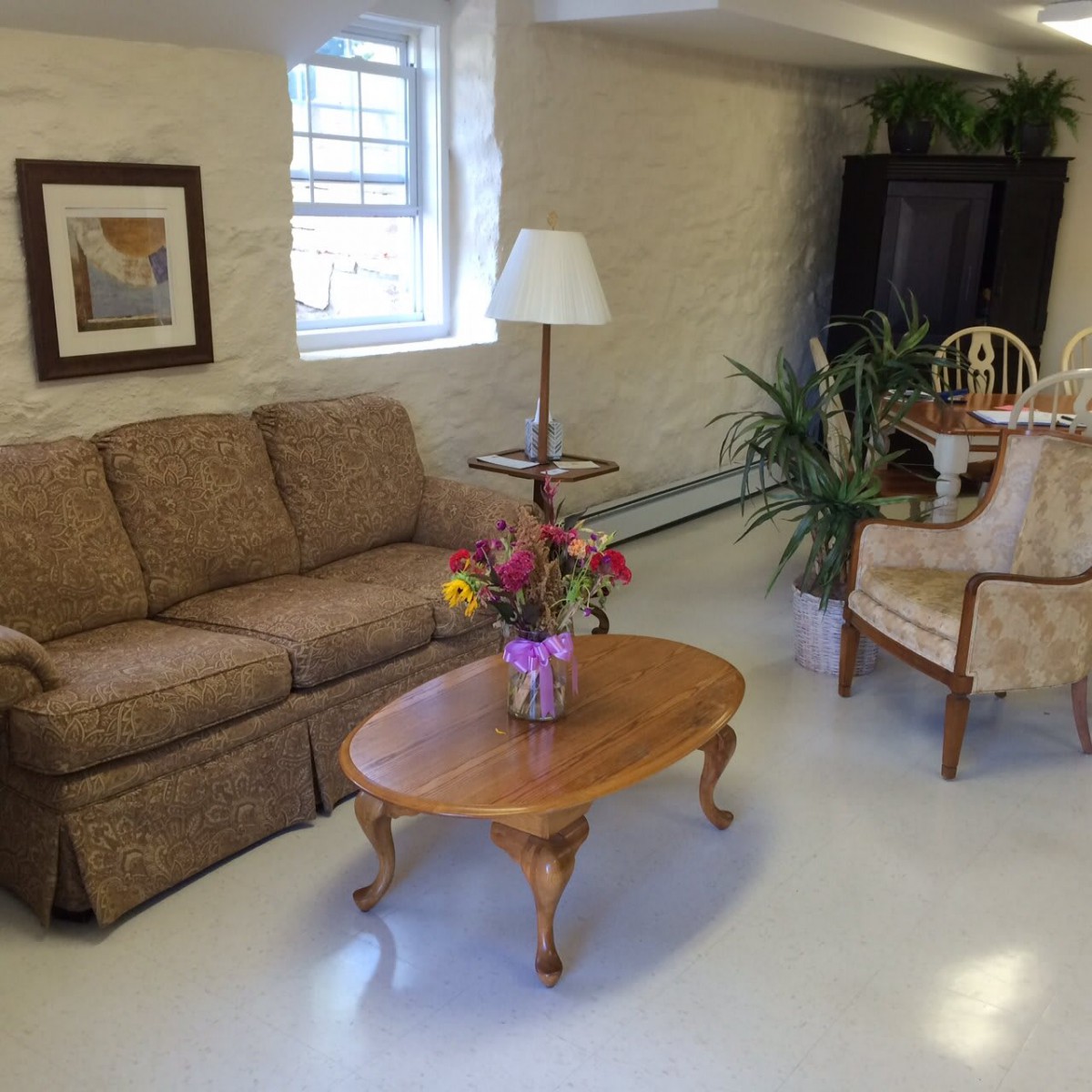 A cozy living room with a sofa, coffee table, lamp, and chairs, adorned with flowers and artwork.