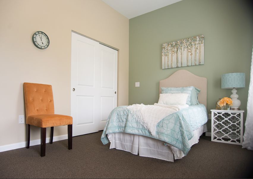 A cozy bedroom featuring a single bed with light blue bedding, an orange chair, and minimalist wall art.