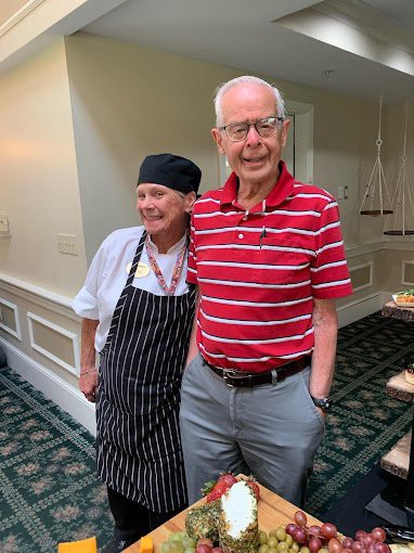 A person in a chef's uniform poses with an elderly man indoors.