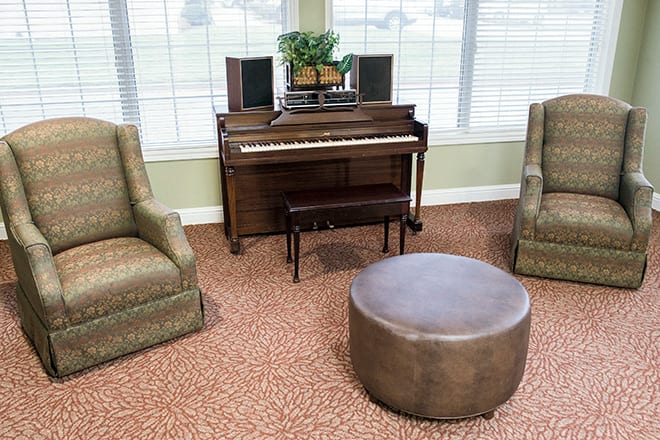 A cozy living room with a piano, two chairs, and a round ottoman.