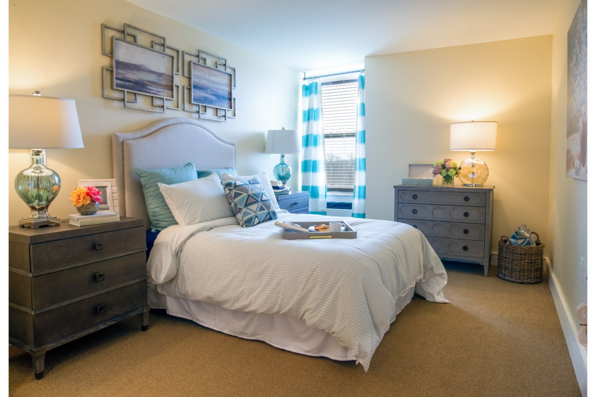 A cozy bedroom featuring a neatly made bed with decorative pillows, flanked by two nightstands and lamps. The room is softly lit and has a window with patterned curtains.