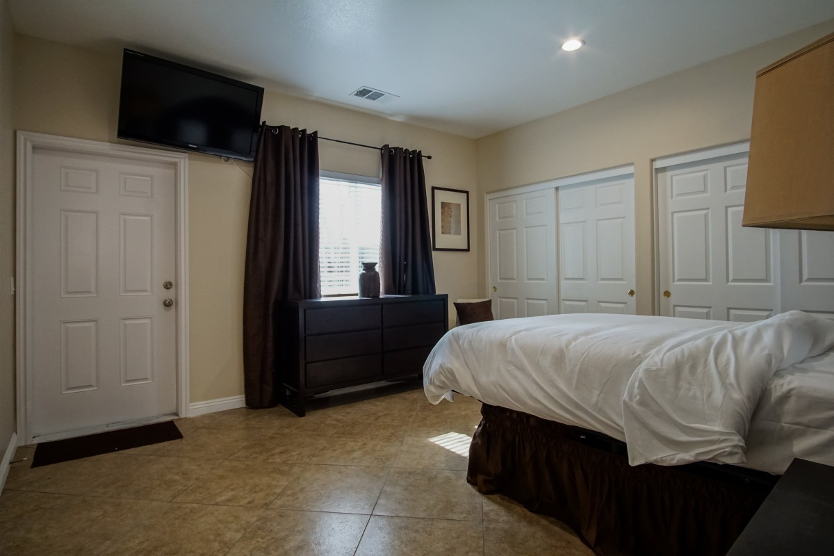 A modern bedroom featuring a neatly made bed, wall-mounted television, and large windows with dark curtains.