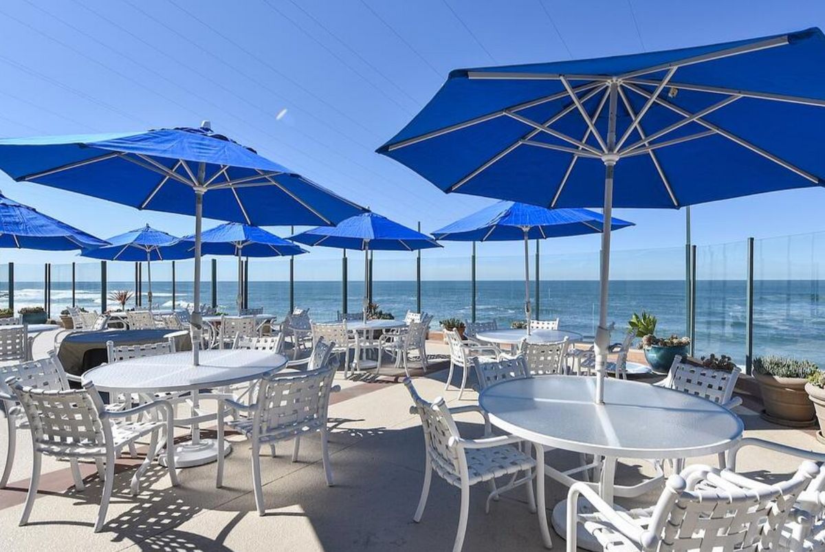 An outdoor beachside restaurant with blue umbrellas and ocean views on a sunny day.