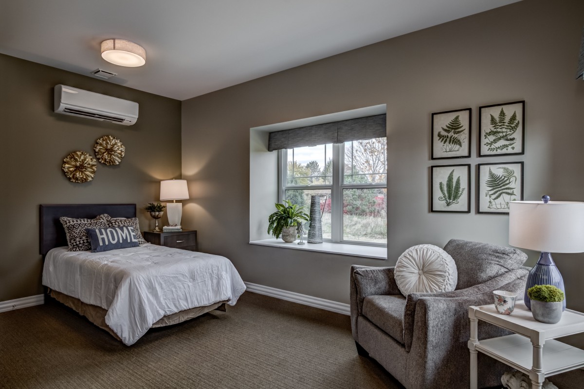 A modern bedroom with minimalist decor, featuring a cozy bed, armchair, and ample natural light from a large window.