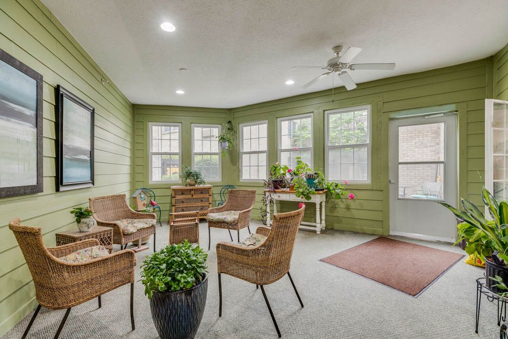 A cozy sunroom featuring wicker chairs, green walls, and plenty of natural light from the large windows.