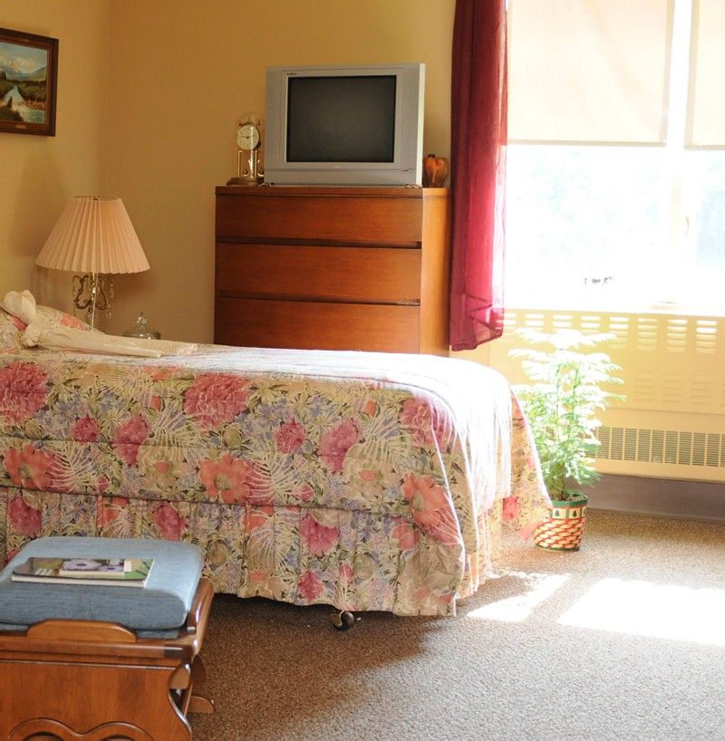 A cozy bedroom featuring a floral-patterned bed, dresser with a television on top, and a lamp by the window.