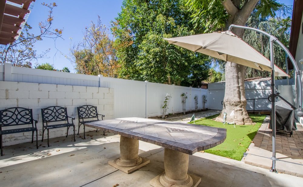 A sunny backyard patio with a stone table, chairs, and an umbrella next to a white fence and trees.