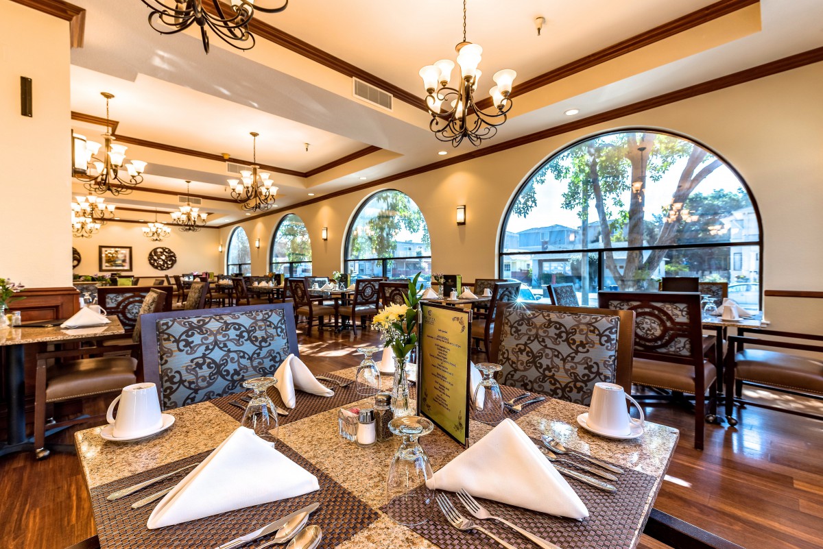 An elegant restaurant dining area with sophisticated table settings and decorative chandeliers, featuring large arched windows that offer a view of the outdoors.