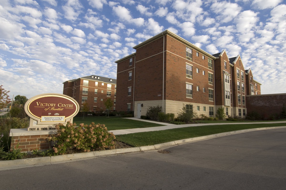 A residential apartment building under a partly cloudy sky with well-kept landscaping and a visible entrance sign.