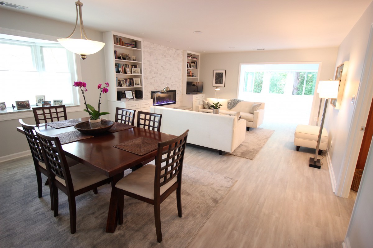An open-concept living and dining area with modern interior design, featuring a wooden dining table, white sofas, and natural light.