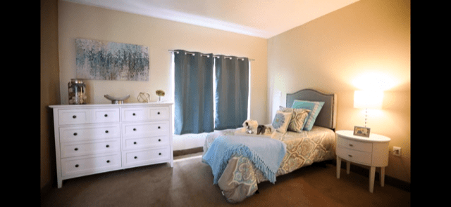 A cozy bedroom with blue curtains and a neatly made single bed, adorned with decorative pillows and soft textures.
